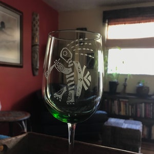 May include: A green wine glass with a decorative etching of a stylized figure playing a musical instrument. The glass has a beaded charm hanging from the stem.