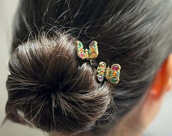 Puede incluir: Dos pequeñas pinzas para el cabello de metal dorado con forma de mariposa, cada una adornada con una variedad de piedras preciosas coloridas de diversas formas en tonos rojo, naranja, amarillo, verde, azul y morado, sujetas en cabello castaño.