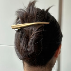 Op de afbeelding: Een gouden haarspeld met een lange, gebogen vorm. De speld houdt het donkerbruine haar van een vrouw in een knot.