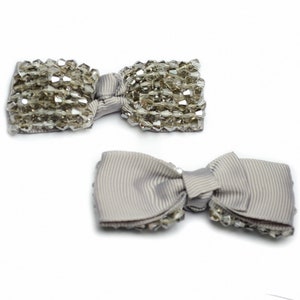 以下が含まれることがあります： クリスタル装飾が施されたグレーの布製のボウが2つ。ボウはシンプルな結び目で結ばれており、わずかにテクスチャのある表面をしています。