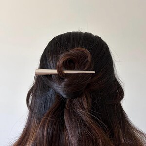 Peut inclure: Un b&acirc;tonnet &agrave; cheveux beige clair, lisse et mat, dot&eacute; d'une extr&eacute;mit&eacute; pointue et effil&eacute;e. L'accessoire est ins&eacute;r&eacute; horizontalement dans une section de cheveux brun fonc&eacute; coiff&eacute;s en un chignon l&acirc;che et rassembl&eacute;, maintenant les cheveux.