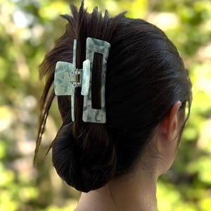 Peut inclure: Grosse pince à cheveux vert clair marbré, fixant des cheveux foncés en chignon. La pince est en matériau translucide, de forme rectangulaire et dotée d'un mécanisme à ressort. L'arrière-plan est une vue floue de feuillage vert.
