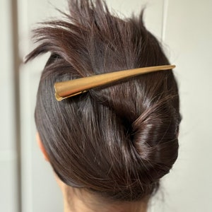 Peut inclure: Une longue pince &agrave; cheveux en m&eacute;tal dor&eacute;, fine et effil&eacute;e, pr&eacute;sentant une forme conique lisse. L'accessoire de coiffure est dot&eacute; d'un m&eacute;canisme &agrave; charni&egrave;re &agrave; la base et est plac&eacute; horizontalement sur des cheveux bruns coiff&eacute;s en chignon.