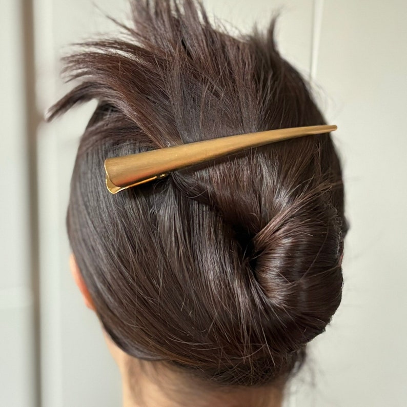 Peut inclure: Une longue pince &agrave; cheveux en m&eacute;tal dor&eacute;, fine et effil&eacute;e, pr&eacute;sentant une forme conique lisse. L'accessoire de coiffure est dot&eacute; d'un m&eacute;canisme &agrave; charni&egrave;re &agrave; la base et est plac&eacute; horizontalement sur des cheveux bruns coiff&eacute;s en chignon.