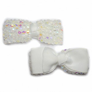 以下が含まれることがあります： 白いヘアボウが2つ。1つのボウは虹色の白いビーズでできており、もう1つは白いサテンリボンでできています。