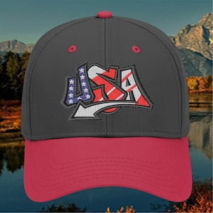 May include: A black and red baseball cap with an embroidered USA graphic in red, white, and blue. The graphic features a stylized USA lettering with stars and stripes.