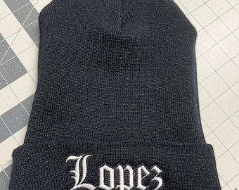 May include: Black knitted beanie with a white embroidered inscription that reads "Lopez" in a stylised font.