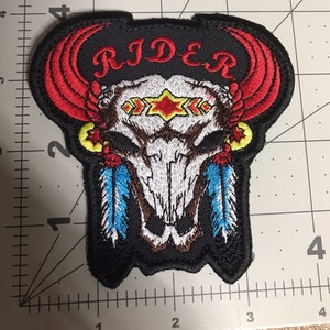 May include: Embroidered patch featuring a white bull skull with red horns, blue feathers, and yellow accents. The word "RIDER" is stitched in red above the skull.