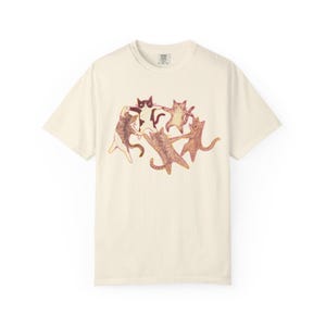 May include: A cream-colored short-sleeve t-shirt featuring a central graphic illustration of five cats in various shades of brown, tan, and black, holding paws and dancing in a circle. The cats have detailed fur textures and expressive faces.