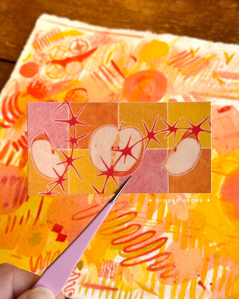 以下が含まれることがあります： リンゴのイラストとスターバーストが描かれた長方形のカラフルな抽象芸術作品。背景はオレンジ、黄色、ピンクの混合色。ピンクのピンセットが前景にあります。