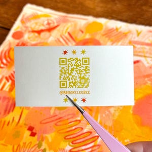 以下が含まれることがあります： 金色のQRコードと「@brookeleebee」のテキストが入った白い名刺が、ピンク色のツールで持たれています。 カードは金色の星形で装飾されており、オレンジと黄色の色合いのカラフルな抽象画の上に置かれています。