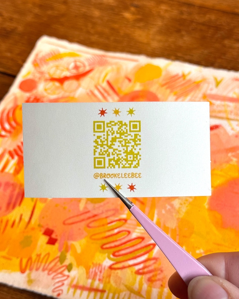 以下が含まれることがあります： 金色のQRコードと「@brookeleebee」のテキストが入った白い名刺が、ピンク色のツールで持たれています。 カードは金色の星形で装飾されており、オレンジと黄色の色合いのカラフルな抽象画の上に置かれています。