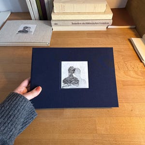 Könnte beinhalten: Ein dunkelblaues Buch mit einem schwarz-weißen abstrakten Bild auf der Vorderseite. Im Hintergrund sind mehrere andere Bücher gestapelt, darunter ein helles Buch mit dem Titel "LA LIBRETTE DE COLETTE".