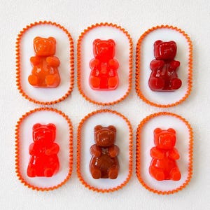 May include: Six oval-shaped white plaques with orange scalloped edges, each featuring a single translucent gummy bear figurine in varying shades of orange, red, dark red, bright red, brown, and light orange, arranged in two rows of three.