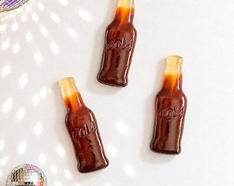 May include: Three cola-flavored gummy candies shaped like glass bottles. Each gummy features a translucent amber neck and a dark brown body with the word cola embossed on the front. A disco ball is visible in the upper left and lower left corners.