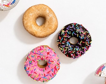 Puede incluir: Tres decoraciones de pared circulares con forma de dónut: un dónut glaseado de color tostado, un dónut con cobertura de chocolate y virutas multicolores, y un dónut con cobertura rosa y virutas multicolores, junto a dos adornos de bola de discoteca.