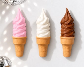 May include: Three wall-mounted ice cream cone sculptures arranged in a row. The left cone features pink soft-serve, the center cone features white soft-serve, and the right cone features brown chocolate soft-serve, all set in tan waffle cones.