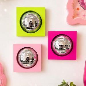 May include: Three square wall-mounted frames in lime green, hot pink, and light pink, each containing a reflective silver disco ball. The disco balls are composed of small mirrored tiles that catch and reflect light, creating a bright, multi-faceted effect.
