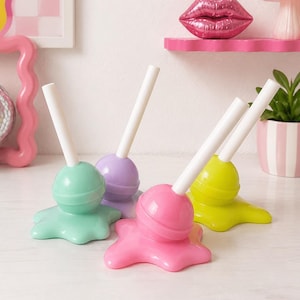 May include: Four decorative lollipop-shaped ornaments in mint green, lavender, bright pink, and lime green. Each piece features a spherical top with a white stick, resting on a base that mimics a melted puddle of candy in a glossy finish.