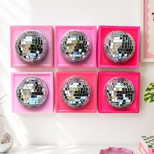 May include: Six square wall art pieces, each featuring a centered silver mirrored disco ball sphere. The frames are painted in varying shades of pink, ranging from light to hot pink. The arrangement is a two-by-three grid on a white wall.
