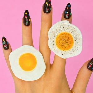 May include: Two fried egg rings featuring a white egg white and a yellow yolk, with one egg showing a speckled black pepper texture. The rings are worn on two fingers, and the fingernails are painted black with colorful polka dot accents.