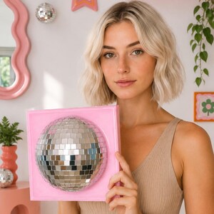 May include: A square, light pink frame featuring a centered, spherical disco ball made of small, reflective silver mirror tiles. The disco ball is embedded within the frame, creating a three-dimensional effect with a smooth, glossy finish.