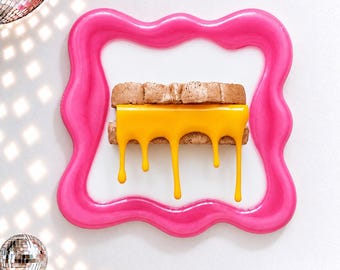 May include: A wall-mounted art piece featuring a grilled cheese sandwich with bright yellow melted cheese dripping down, encased in a glossy, wavy, hot pink frame. The sandwich has a textured, toasted brown bread appearance.