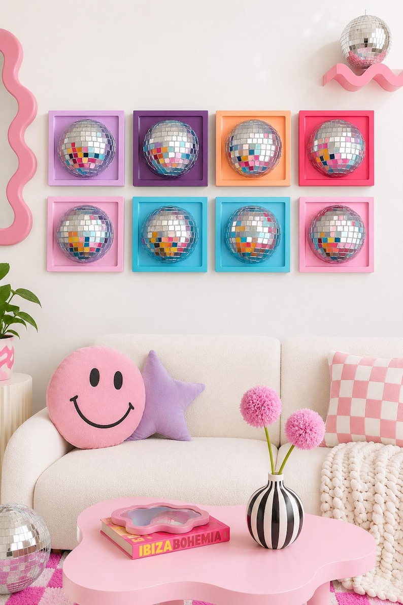 May include: Eight square wall frames in purple, orange, pink, and blue, each containing a reflective disco ball. Below, a pink smiley face pillow, a purple star pillow, a pink checkered pillow, and a black and white striped vase with pink flowers.