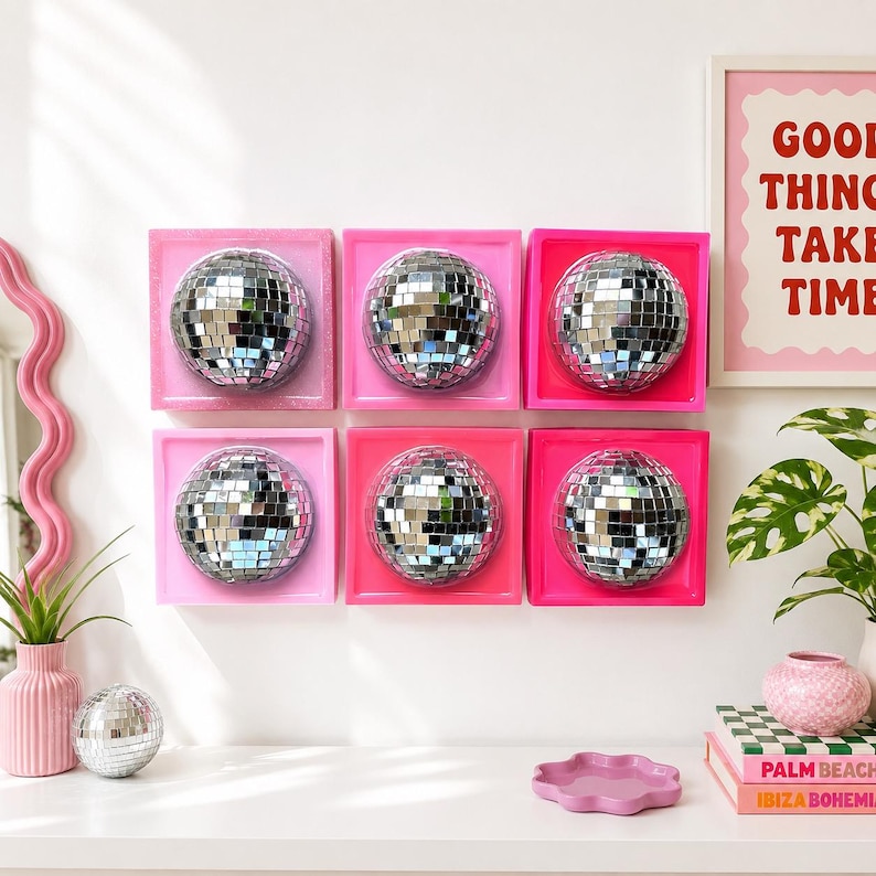 May include: Six square wall art pieces, each featuring a centered silver mirrored disco ball sphere. The frames are painted in varying shades of pink, ranging from light to hot pink. The arrangement is a two-by-three grid on a white wall.