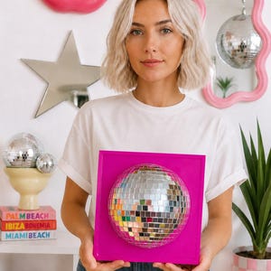 Pode incluir: Uma moldura de parede quadrada em rosa brilhante com uma bola de discoteca esf&eacute;rica centralizada, feita de pequenos azulejos espelhados refletores. A bola de discoteca est&aacute; fixada na base quadrada rosa, criando uma pe&ccedil;a tridimensional.