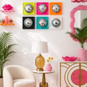 May include: A wall display featuring six square frames with mirrored disco balls, a pink lip sculpture, two wavy shelves, a cream armchair, a gold lamp with a white shade, a pink vase, and a white cabinet with gold geometric accents.