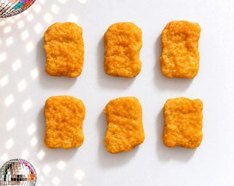 May include: Six golden-brown, breaded chicken nuggets arranged in two rows of three, accompanied by two small, reflective disco balls positioned in the top-left and bottom-left corners of the frame, casting light patterns across the surface.