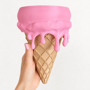 May include: A decorative container shaped like an ice cream cone with a tan, waffle-textured base and a smooth, bright pink top section featuring realistic drips cascading down the sides of the cone. The object has a wide, open circular top.