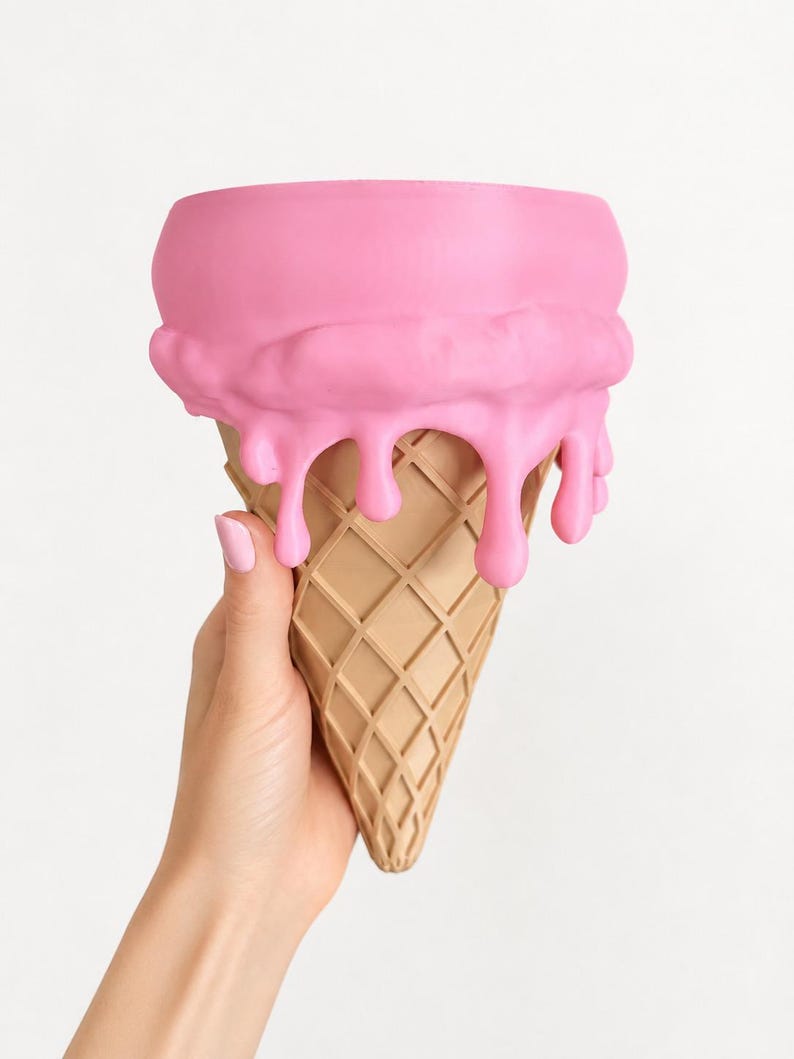 May include: A decorative container shaped like an ice cream cone with a tan, waffle-textured base and a smooth, bright pink top section featuring realistic drips cascading down the sides of the cone. The object has a wide, open circular top.