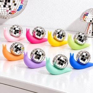 May include: Eight snail-shaped figurines in various colors including pink, hot pink, yellow, lime green, orange, purple, mint, and blue. Each snail features a reflective, mirrored disco ball shell made of small square tiles.
