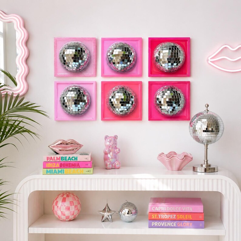 May include: Six square wall art pieces featuring silver disco balls in varying shades of pink frames. A silver disco ball lamp, a pink gummy bear figurine, a pink lip-shaped dish, a checkered sphere, a star, and stacked books on a white console.