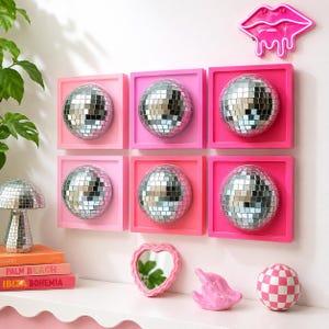 May include: Six square wall-mounted frames in varying shades of pink, each containing a silver mirrored disco ball sphere. A neon pink lip-shaped light, a mushroom-shaped disco ball, a heart-shaped mirror, a pink sculpture, and a checkered ball.