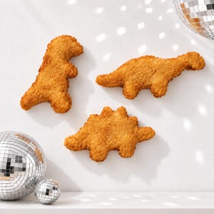 May include: Three golden-brown, breaded dinosaur-shaped food items arranged on a white surface. The shapes resemble a standing T-Rex, a long-necked Brontosaurus, and a Stegosaurus. Two silver mirrored disco balls are positioned in the corners.