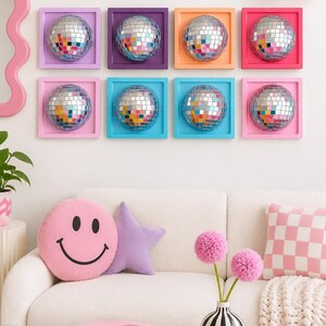 Pode incluir: Oito molduras de parede quadradas em roxo, laranja, rosa e azul, cada uma contendo uma bola de discoteca refletora. Abaixo, uma almofada rosa com rosto sorridente, uma estrela roxa, uma almofada xadrez e o livro IBIZA BOHEMIA numa mesa rosa.