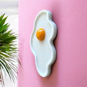 Puede incluir: Un objeto decorativo de pared de cer&aacute;mica con forma de huevo frito, que presenta una base ovalada blanca y lisa con una yema amarilla redondeada, brillante y elevada en el centro, con un contorno org&aacute;nico irregular.