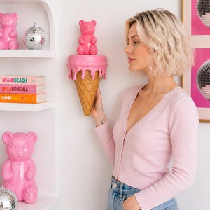 May include: A wall-mounted shelf shaped like an ice cream cone with a pink melting top and a tan waffle cone base. A glossy pink gummy bear figurine sits on the flat surface of the shelf. The shelf is mounted on a white wall near white shelves.