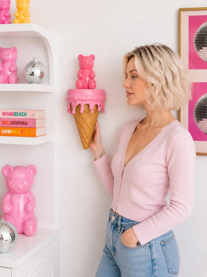 May include: A wall-mounted shelf shaped like an ice cream cone with a pink melting top and a tan waffle cone base. A glossy pink gummy bear figurine sits on the flat surface of the shelf. The shelf is mounted on a white wall near white shelves.