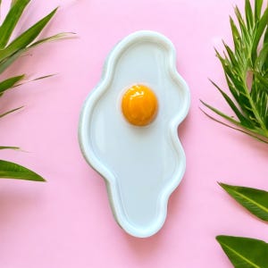 Puede incluir: Un plato de cer&aacute;mica blanca con forma de huevo frito y contorno irregular y ondulado. El centro presenta una yema circular brillante, elevada y de color naranja intenso. El plato tiene una superficie lisa y reflectante sobre un fondo rosa.