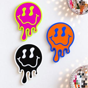 May include: Three wall-mounted smiley face signs with a melting effect. One sign is neon pink with a lime green outline, one is orange with a blue outline, and one is black with a white outline. Each features distorted, elongated eyes and a wavy mouth.