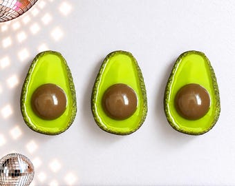 Puede incluir: Tres mitades de aguacate montadas en la pared con pulpa de color verde lima brillante y un hueso marrón liso y brillante en el centro. Cada pieza tiene una corteza exterior verde oscuro texturizada y un acabado pulido que refleja la luz.
