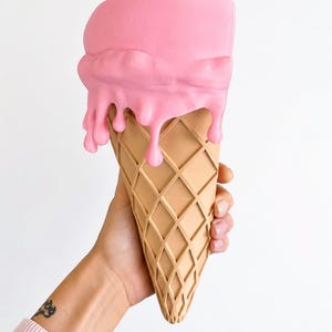 May include: A wall-mounted planter shaped like an ice cream cone with a tan, waffle-textured base and a smooth, matte pink scoop on top featuring realistic drips cascading down the sides of the cone, creating a functional decorative container.