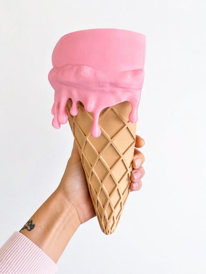 May include: A wall-mounted planter shaped like an ice cream cone with a tan, waffle-textured base and a smooth, matte pink scoop on top featuring realistic drips cascading down the sides of the cone, creating a functional decorative container.