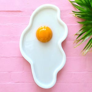Puede incluir: Una bandeja de cer&aacute;mica blanca con forma de huevo frito y contorno irregular y org&aacute;nico. El centro presenta un elemento circular elevado, brillante y de color naranja vivo que representa la yema, sobre una base blanca c&oacute;ncava.