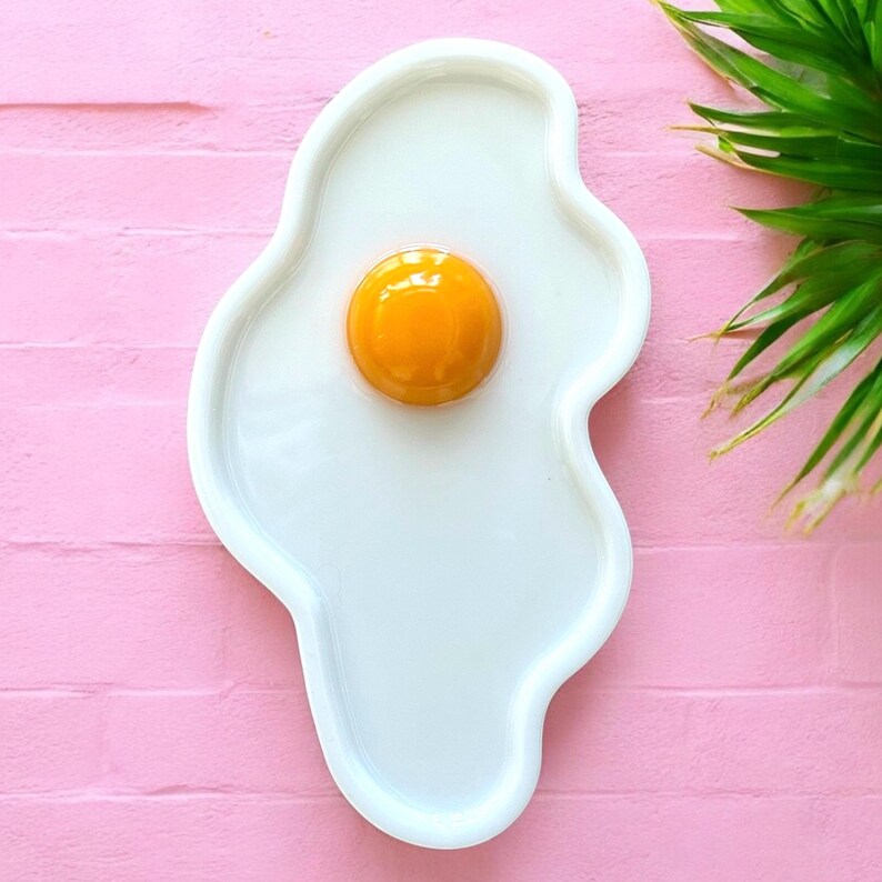Puede incluir: Una bandeja de cer&aacute;mica blanca con forma de huevo frito y contorno irregular y org&aacute;nico. El centro presenta un elemento circular elevado, brillante y de color naranja vivo que representa la yema, sobre una base blanca c&oacute;ncava.