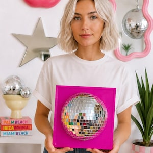 May include: A square, bright pink wall-mounted frame featuring a centered, spherical disco ball made of small, reflective silver mirror tiles. The disco ball is securely attached to the flat, solid pink square base, creating a dimensional effect.